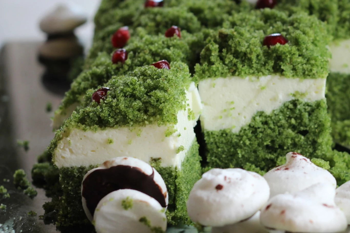 Deser z zielonym biszkoptem, warstwą jasnego kremu i drobnymi czerwonymi owocami na wierzchu, wokół znajdują się białe bezy z ciemną polewą.