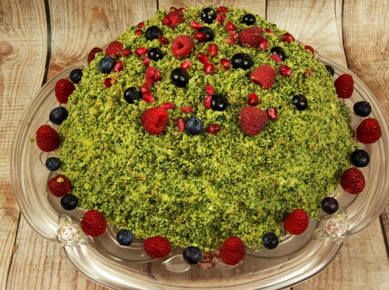 Ciasto o kopułowym kształcie pokryte intensywnie zieloną kruszonką, udekorowane świeżymi malinami, borówkami oraz ziarnami granatu, ułożone na przezroczystej paterze na tle drewnianych desek.