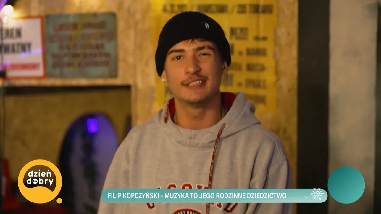 Mężczyzna w jasnej bluzie z kapturem oraz czarnej czapce na głowie, stojący na tle przyciemnionego wnętrza z kolorowymi tablicami i światłami. Na dole ekranu pasek z napisem informacyjnym od programu telewizyjnego.