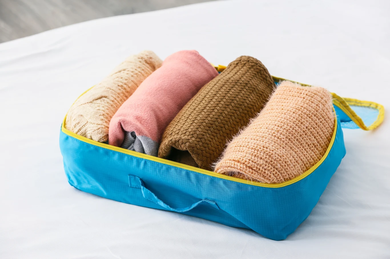 Nigdy nie wkładam ubrań bezpośrednio do walizki, zawsze używam organizerów Niebieska torba z żółtym zamkiem błyskawicznym, w której znajdują się zrolowane swetry w różnych kolorach – beżowym, różowym, brązowym oraz jasnobeżowym, umieszczone na białym tle.