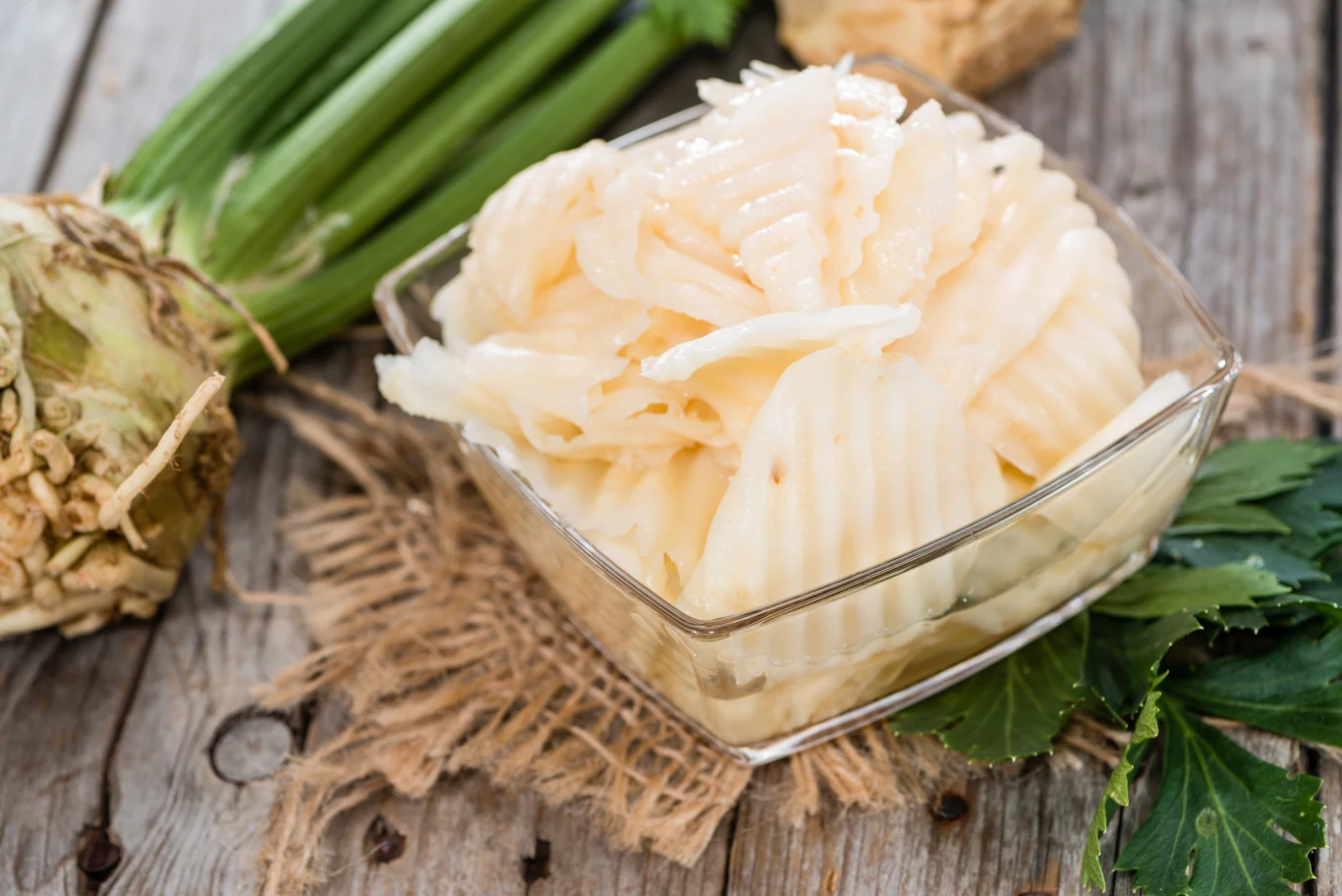 Pokrojony w faliste plasterki seler umieszczony w szklanej miseczce, obok świeża bulwa selera z natką oraz zielone liście, całość na drewnianym stole z jutową podkładką.