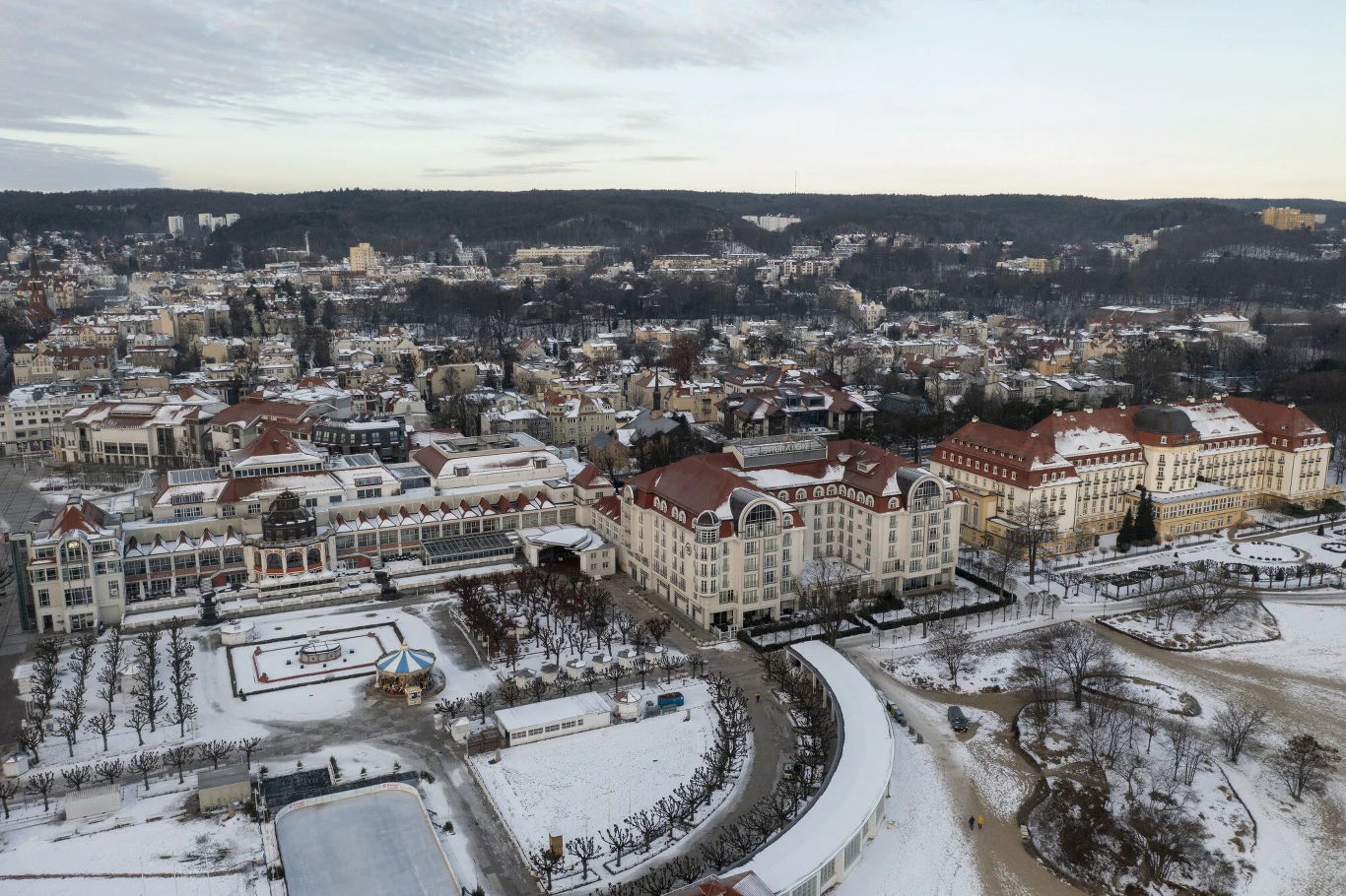 Za nocleg w sylwestra w Sopocie trzeba zapłacić ponad 500 zł Panorama miasta w zimowej scenerii z zabytkowym, dużym budynkiem o czerwonym dachu, otoczonym mniejszymi budowlami oraz drzewami pokrytymi śniegiem; w oddali widać las i pagórkowaty teren.