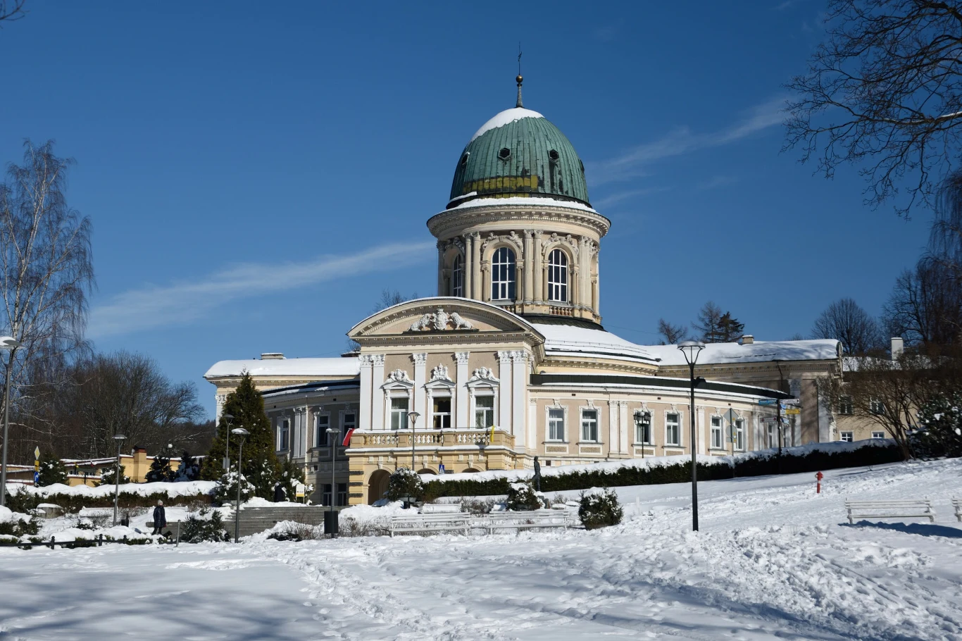 Lądek-Zdrój z powodzeniem wykorzystuje gminną farmę fotowoltauczną. Na zdj. Zdrój Wojciech w Lądku-Zdroju Klasycystyczny budynek z zieloną kopułą otoczony zaśnieżonym ogrodem i drzewami, po lewej stronie widoczne bezlistne drzewa, na pierwszym planie ślady na śniegu oraz krzewy.