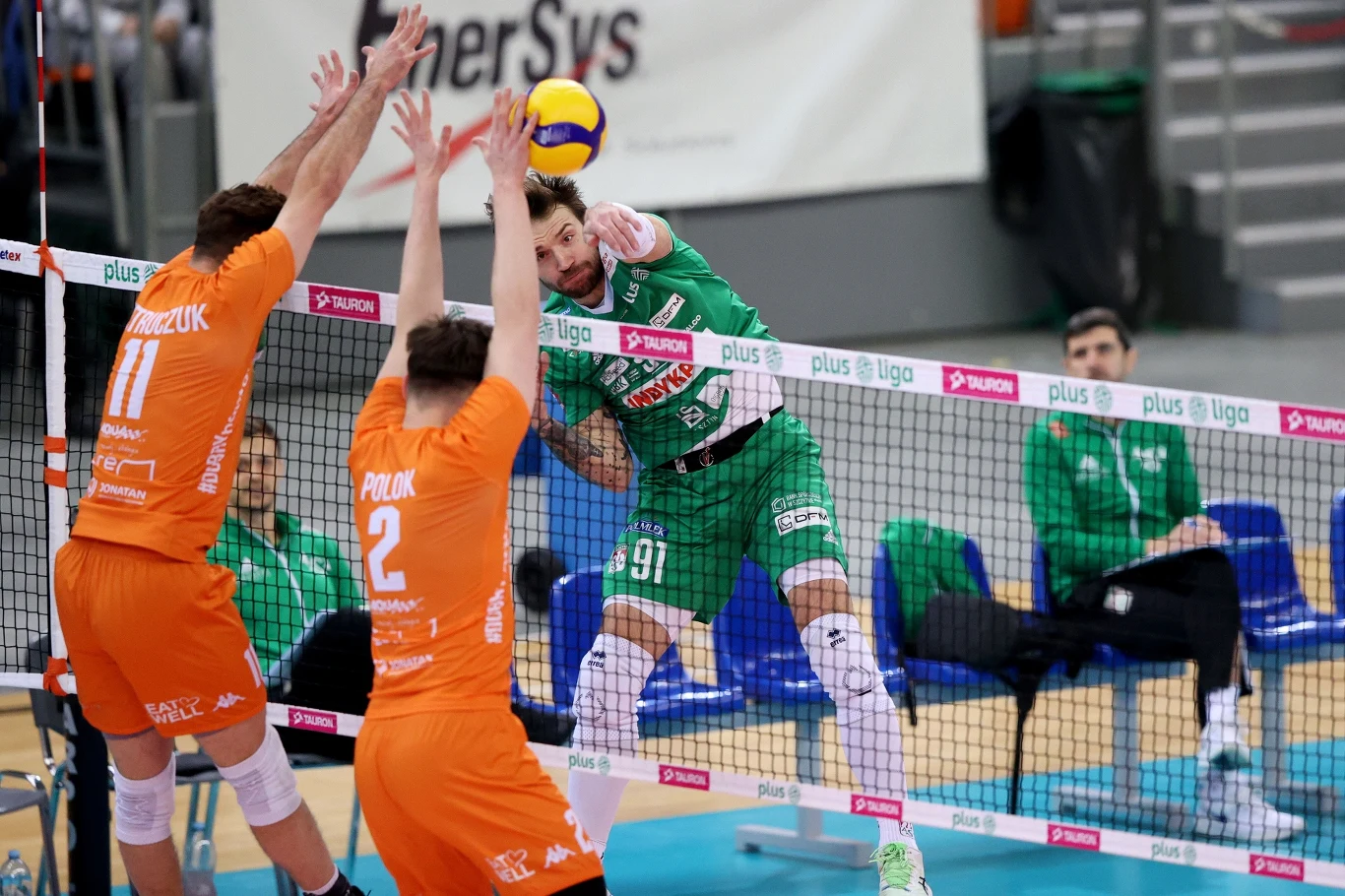 Zawodnik w zielonym stroju atakuje piłkę siatkową nad siatką, podczas gdy dwóch graczy w pomarańczowych strojach próbuje zablokować atak. Drużyny rywalizują podczas meczu siatkówki na hali sportowej.