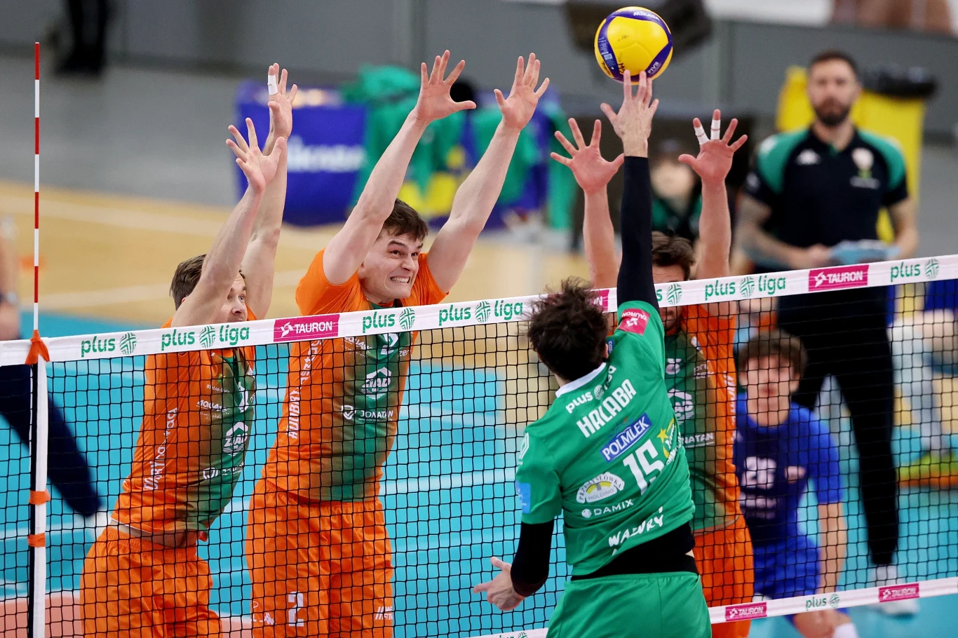 Zawodnik drużyny w zielonym stroju atakuje piłkę siatkową nad siatką, naprzeciwko trzech zawodników w pomarańczowych koszulkach próbujących zablokować atak. W tle widoczni są trener oraz inni zawodnicy.
