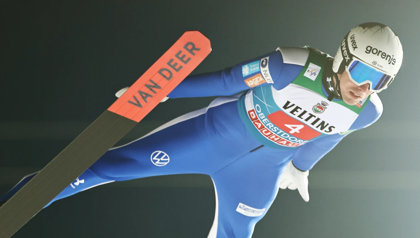 Skoczek narciarski ubrany w niebieski kombinezon i kask ochronny unosi się w powietrzu podczas skoku, widoczne logo sponsora oraz numer startowy, tło rozmyte, akcent na dynamiczny ruch.