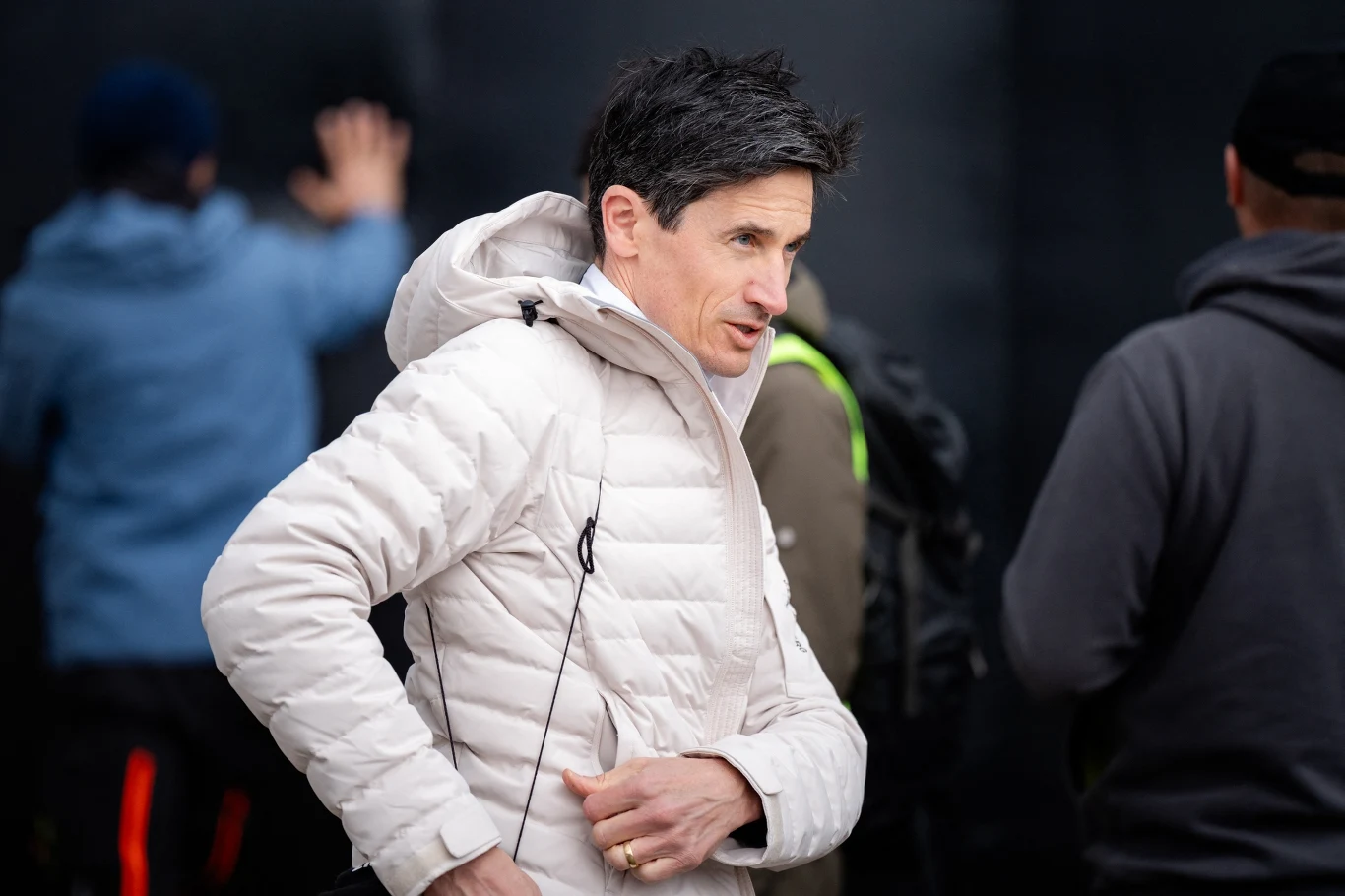 Mężczyzna w jasnej kurtce na pierwszym planie, patrzący w bok, w tle dwie rozmazane postacie w ciemnych ubraniach.