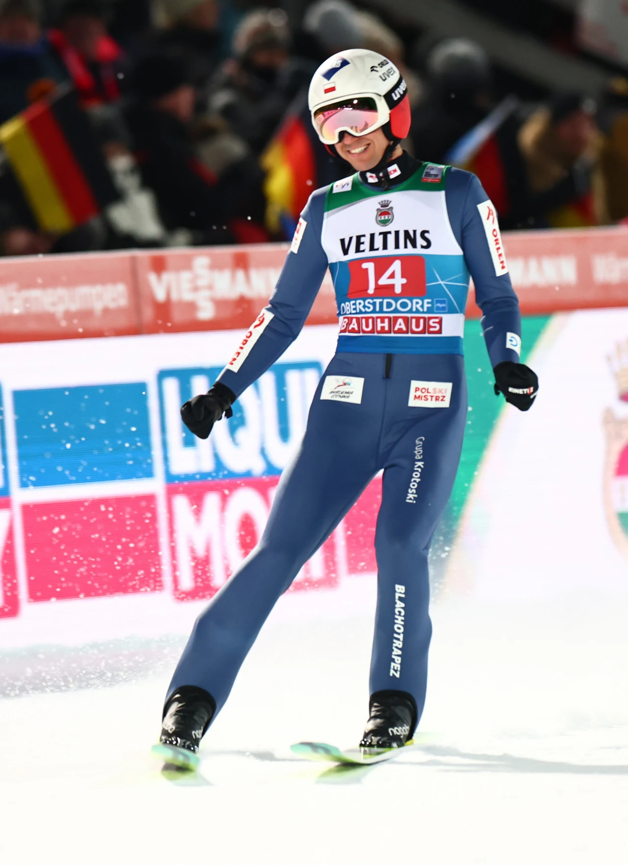 Zawodnik skoków narciarskich w niebieskim kombinezonie i kasku, stojący na nartach na skoczni, z wyraźnym numerem startowym 14, w tle barwne tablice reklamowe i elementy kibicowania publiczności.