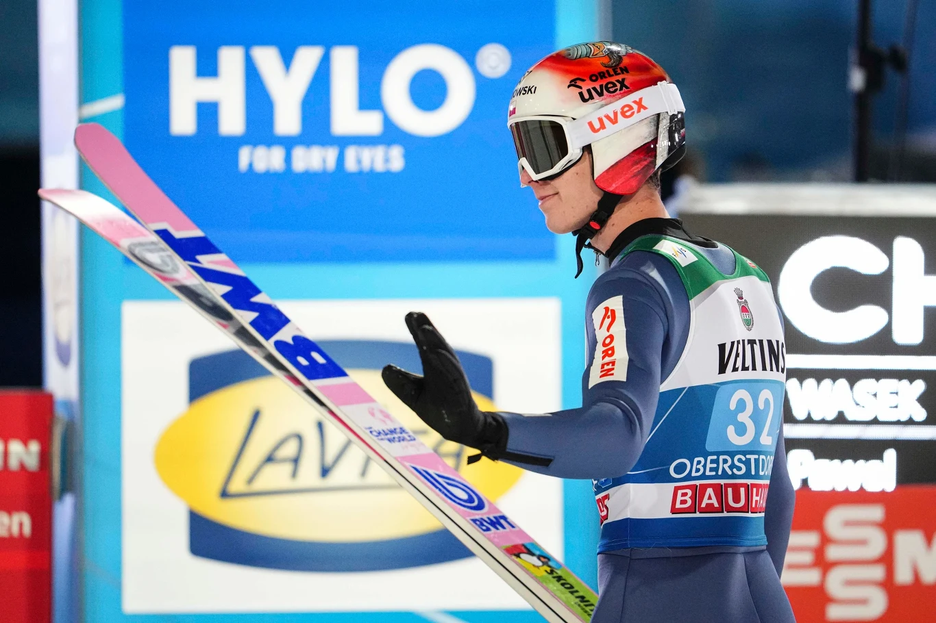Paweł Wąsek Zawodnik w kombinezonie narciarskim i kasku trzyma długą nartę, stojąc na tle reklam sponsorskich podczas zawodów skoków narciarskich.