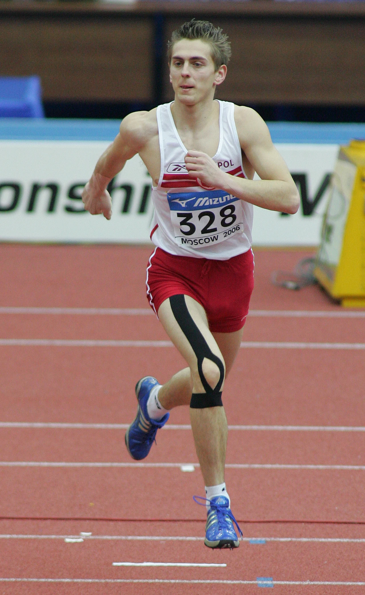 Robert Wolski Lekkoatleta w biegu na hali lekkoatletycznej, ubrany w biało-czerwony strój z numerem startowym 328 oraz opaską kompresyjną na kolanie.