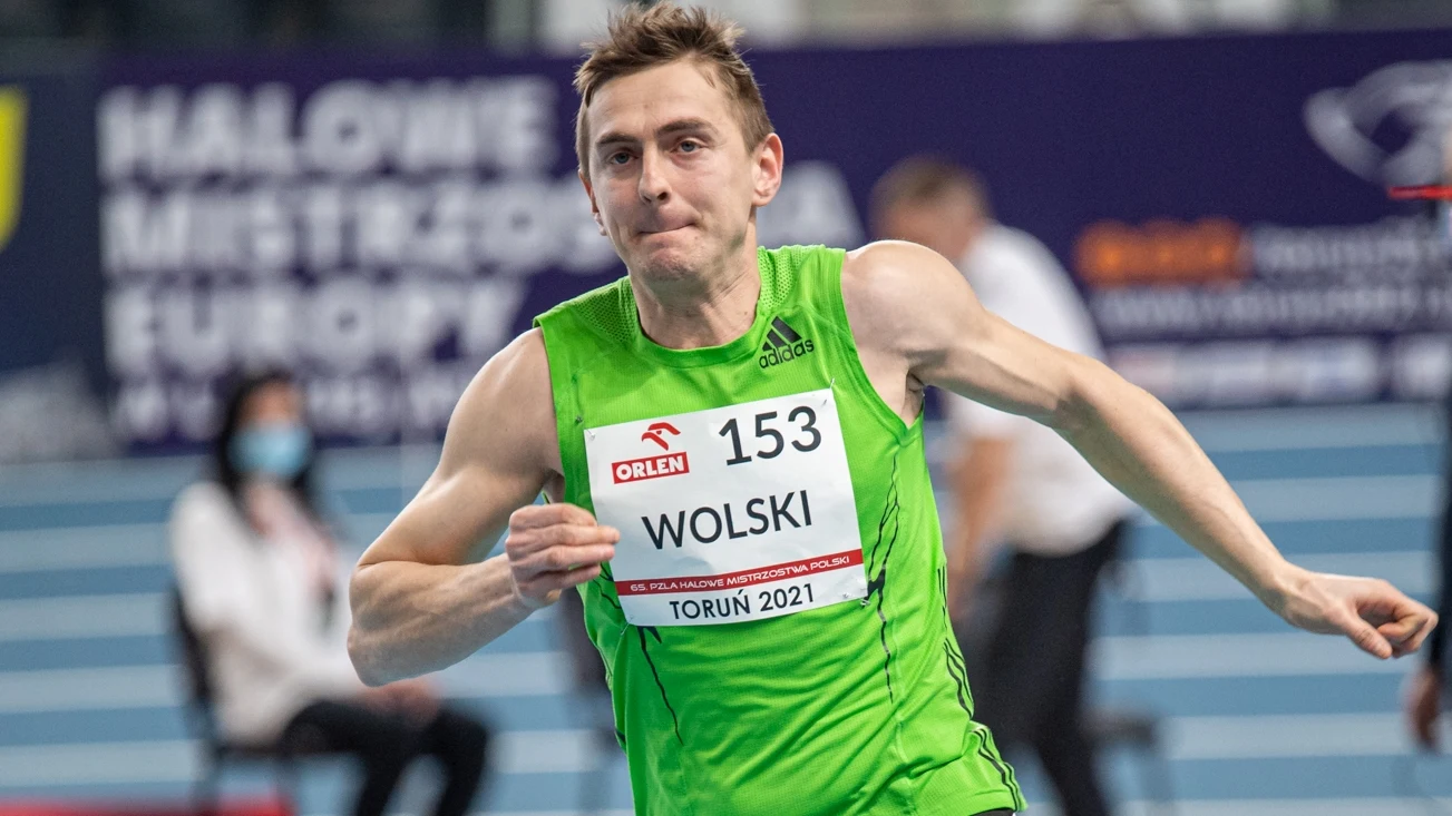 Robert Wolski Lekkoatleta w zielonej koszulce i numerze startowym 153 Wolski podczas biegu na zawodach halowych. W tle bieżnia i rozmazane sylwetki innych osób.