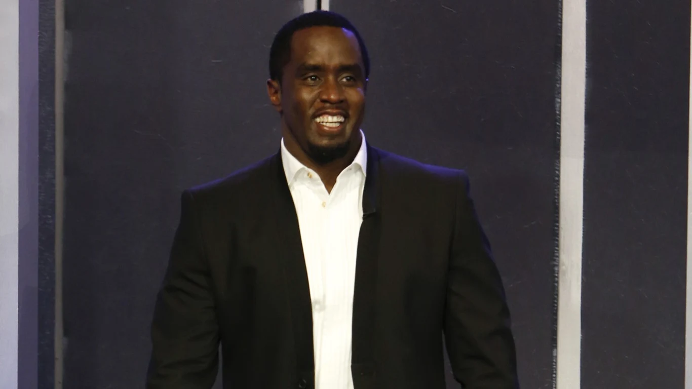 Sean "Diddy" Combs Mężczyzna w eleganckim czarnym garniturze i białej koszuli stoi na tle ciemnej ścianki, uśmiecha się szeroko.