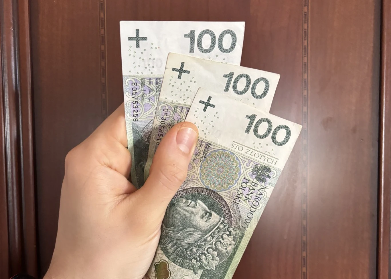 Nawet 300 zł dofinansowania w pracy. Tyle można dostać na wniosek od pracodawcy. Trzy banknoty o nominale 100 złotych trzymane w lewej dłoni na tle drewnianych drzwi.
