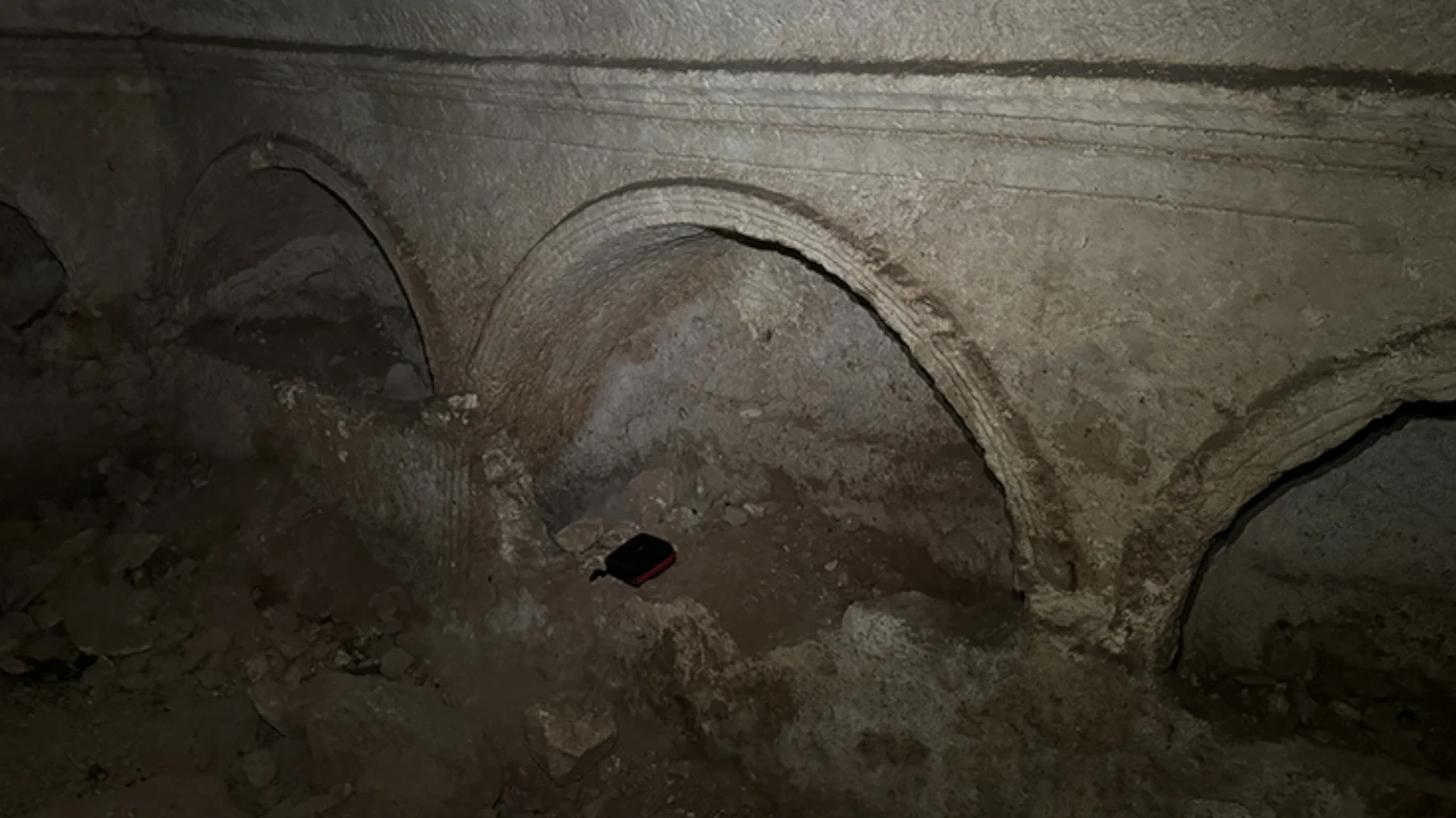 Wnętrze grobowca Stare, zniszczone podziemia z kamiennymi łukami i wyżłobieniami na ścianach. Wśród gruzu i ziemi znajduje się niewielki przedmiot, prawdopodobnie książka, leżący na ziemi.