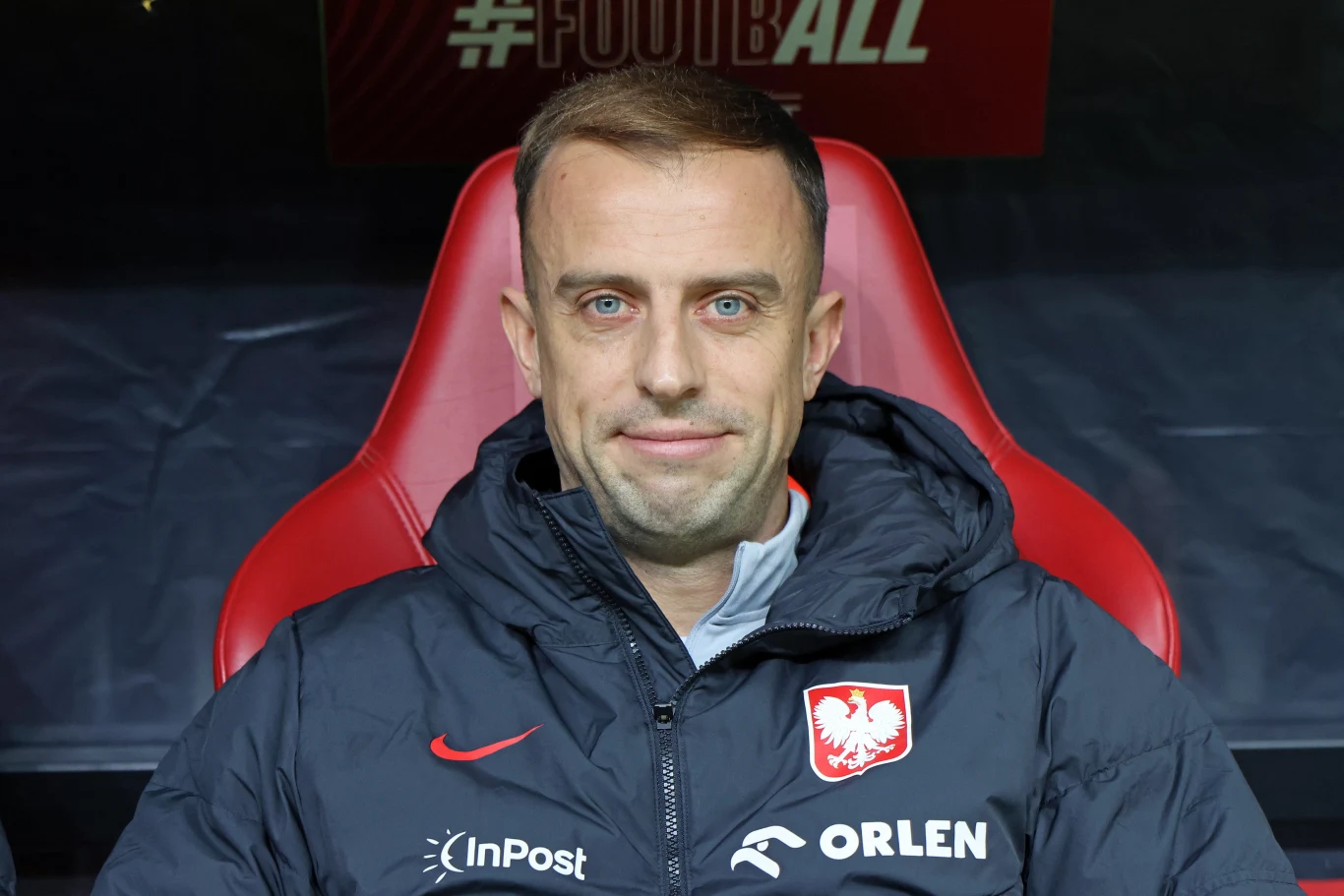Kamil Grosicki Mężczyzna siedzący na stadionowym krześle, ubrany w kurtkę z godłem Polski i logotypami sponsorów, w tle rozmyta część stadionu.