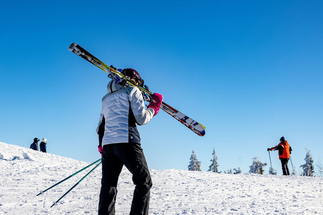 Osoba ubrana w zimowy strój niesie narty na ramieniu i kijki, idąc po ośnieżonym stoku. W tle widać kilka innych osób na śniegu, niebo jest bezchmurne i niebieskie.