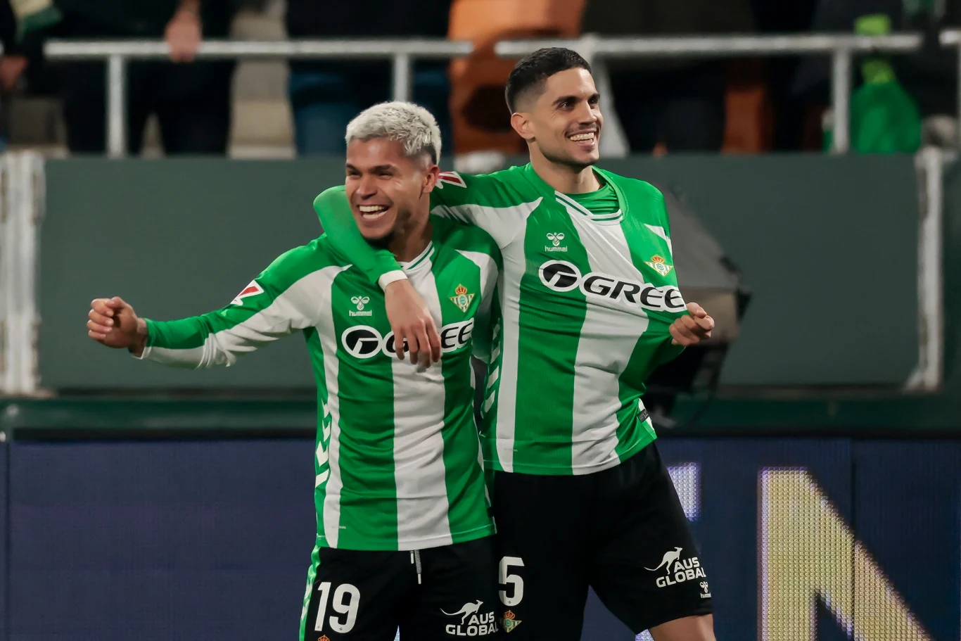 Dwóch piłkarzy w zielono-białych koszulkach drużyny Real Betis celebruje wspólnie sukces na boisku, jeden z zawodników obejmuje drugiego ramieniem, obaj uśmiechnięci, w tle trybuny z kibicami.