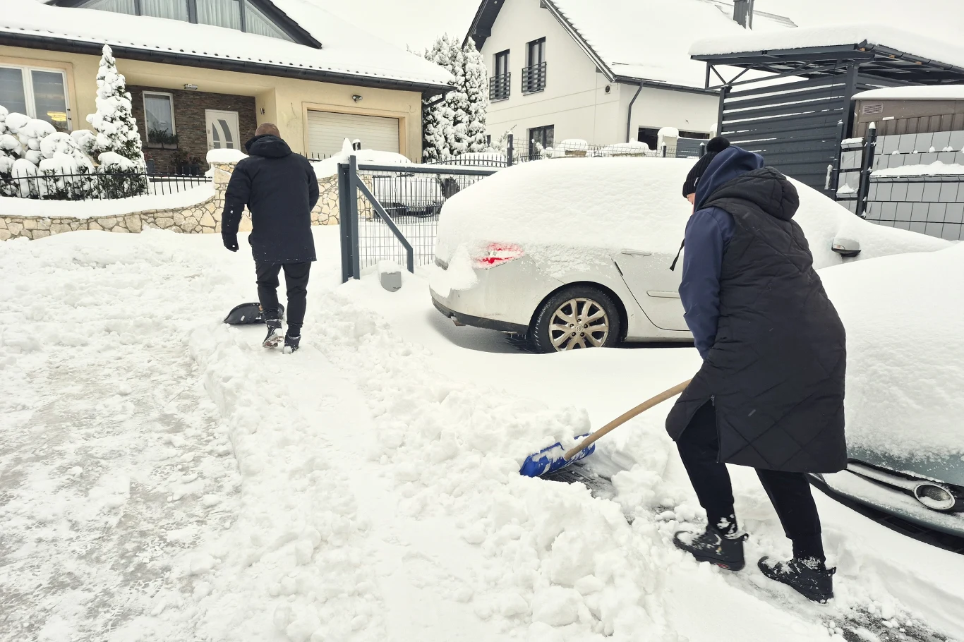 Dwie osoby odśnieżają podjazd i przestrzeń wokół samochodu przed domem w zimowy, śnieżny dzień. Pojazdy i dachy pokryte grubą warstwą śniegu, ogrodzenie oraz domy w tle dodają scenerii charakterystycznej dla spokojnej dzielnicy mieszkalnej.