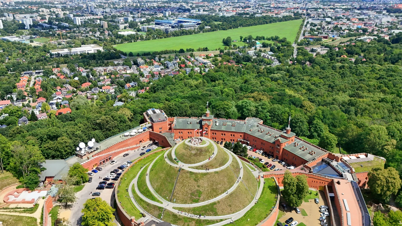 Zabytkowy kopiec otoczony ceglanymi murami twierdzy, położony wśród zieleni, z widokiem na rozległe miasto w tle i okoliczne osiedla mieszkalne.