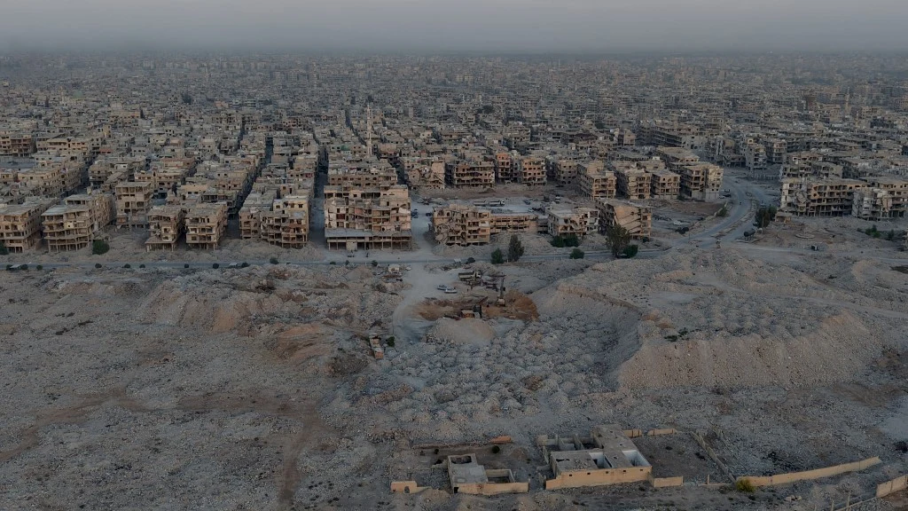 Panorama rozległego, zniszczonego miasta z licznymi opuszczonymi budynkami, gruzowiskami oraz wyjałowioną okolicą bez widocznych ludzi. Szeroki kadr uwydatnia skalę zniszczeń i pustkę po dawnych zabudowaniach.