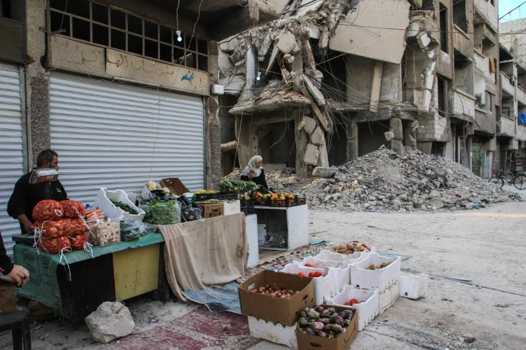 Stoisko z warzywami i owocami rozstawione na ulicy przed zrujnowanym budynkiem, w tle widać gruzowisko i poważnie uszkodzoną fasadę, przy stoisku stoją dwie osoby handlujące produktami.