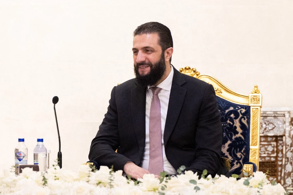 Mężczyzna w garniturze siedzący na ozdobnym, złotym krześle przy stole pokrytym białymi kwiatami, w tle widoczny mikrofon oraz butelki z wodą