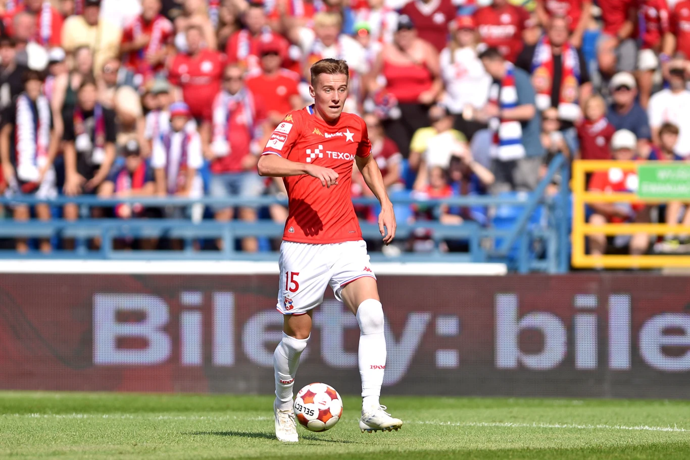 Wiktor Staszak Piłkarz w czerwonej koszulce z numerem 15 prowadzi piłkę po murawie stadionu, w tle trybuny wypełnione kibicami ubranymi w klubowe barwy.