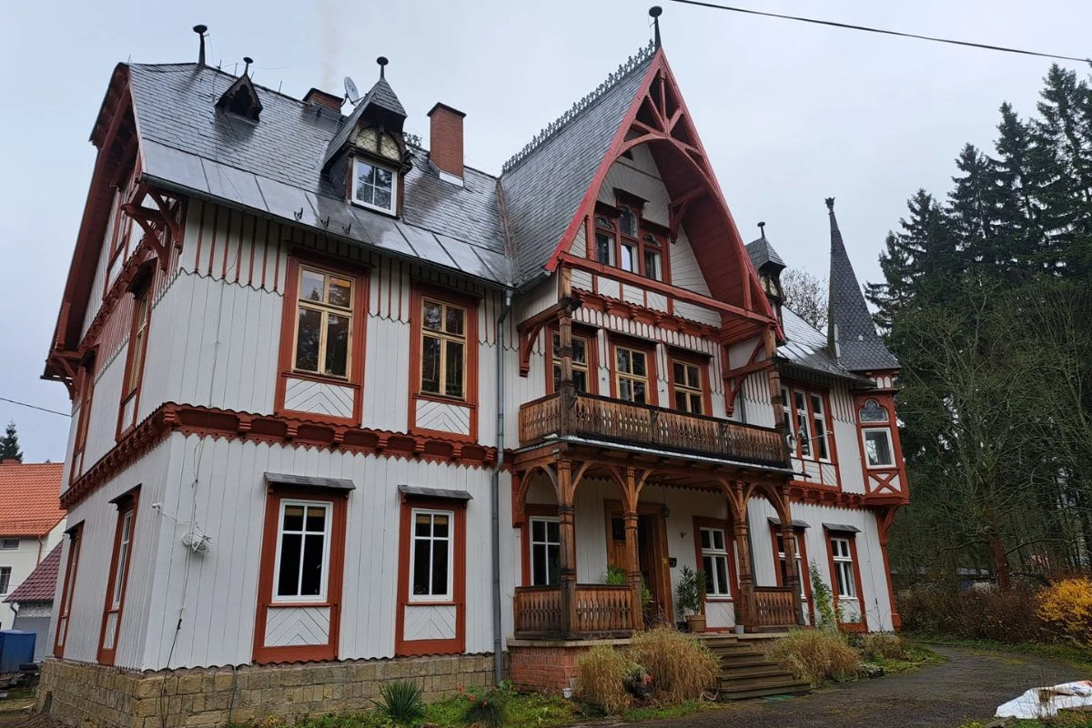 Duży, zabytkowy budynek o bogatej drewnianej konstrukcji i licznych zdobieniach w stylu szwajcarskim, z wysokim, stromym dachem, balkonem oraz rzeźbionymi detalami, otoczony zielenią i innymi domami.