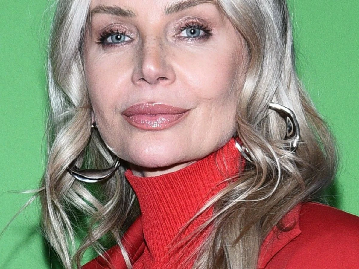 Agnieszka Woźniak-Starak Kobieta o jasnych włosach i niebieskich oczach w czerwonym swetrze, z delikatnym makijażem i dużymi, srebrnymi kolczykami na zielonym tle.