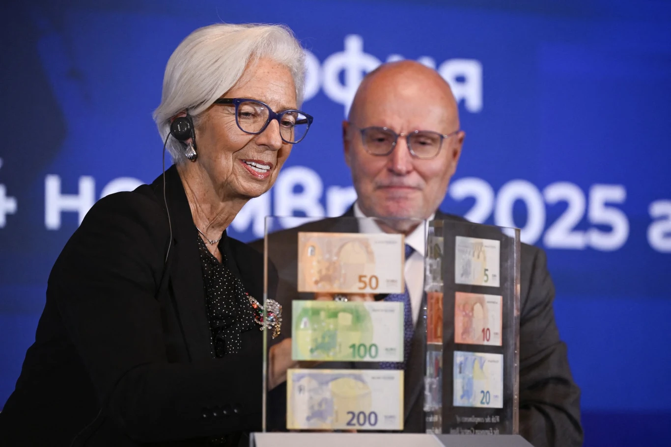 Szefowa Europejskiego Banku Centralnego Christine Lagarde wita Bułgarię w strefie euro Dwie osoby w eleganckich ubraniach stoją przy ekspozytorze z sześcioma banknotami euro o różnych nominałach. Kobieta ze słuchawką na uchu pokazuje banknoty, mężczyzna stoi w tle, oboje uśmiechnięci. W tle widoczny niebieski ekran z białym napisem.