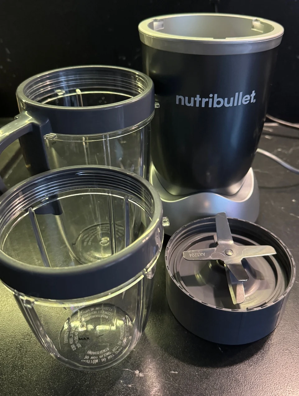 Blender kielichowy marki Nutribullet z zestawem dwóch przezroczystych kubków, nakładką z ostrzami oraz bazą elektryczną, ustawione na ciemnym blacie.