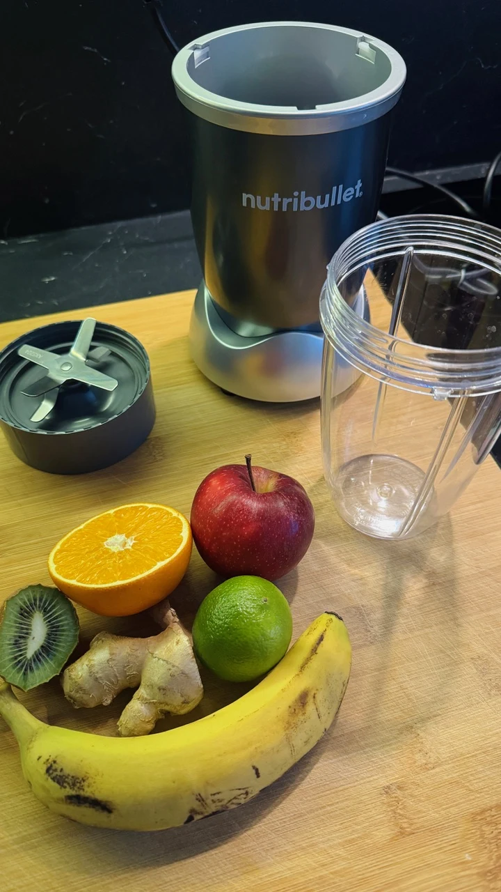Blender marki Nutribullet razem z rozkręconym pojemnikiem oraz owocami i kawałkiem imbiru leżącymi na desce do krojenia, wśród owoców znajdują się pomarańcza, jabłko, kiwi, limonka i banan.