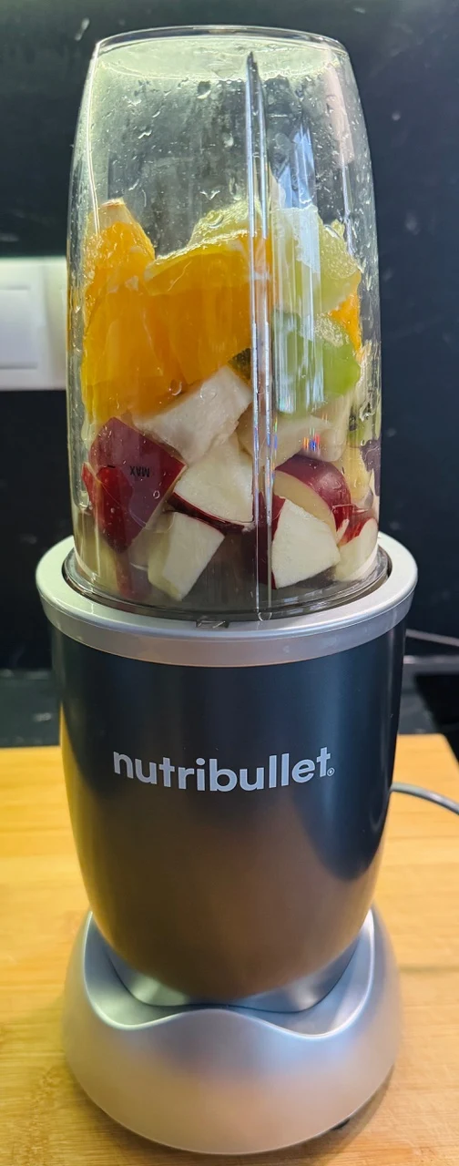 Blender marki Nutribullet wypełniony kawałkami świeżych owoców, takich jak jabłka, pomarańcze i kiwi, stoi na drewnianym blacie kuchennym. Tło stanowi ciemna ściana z widocznym fragmentem gniazdka elektrycznego.
