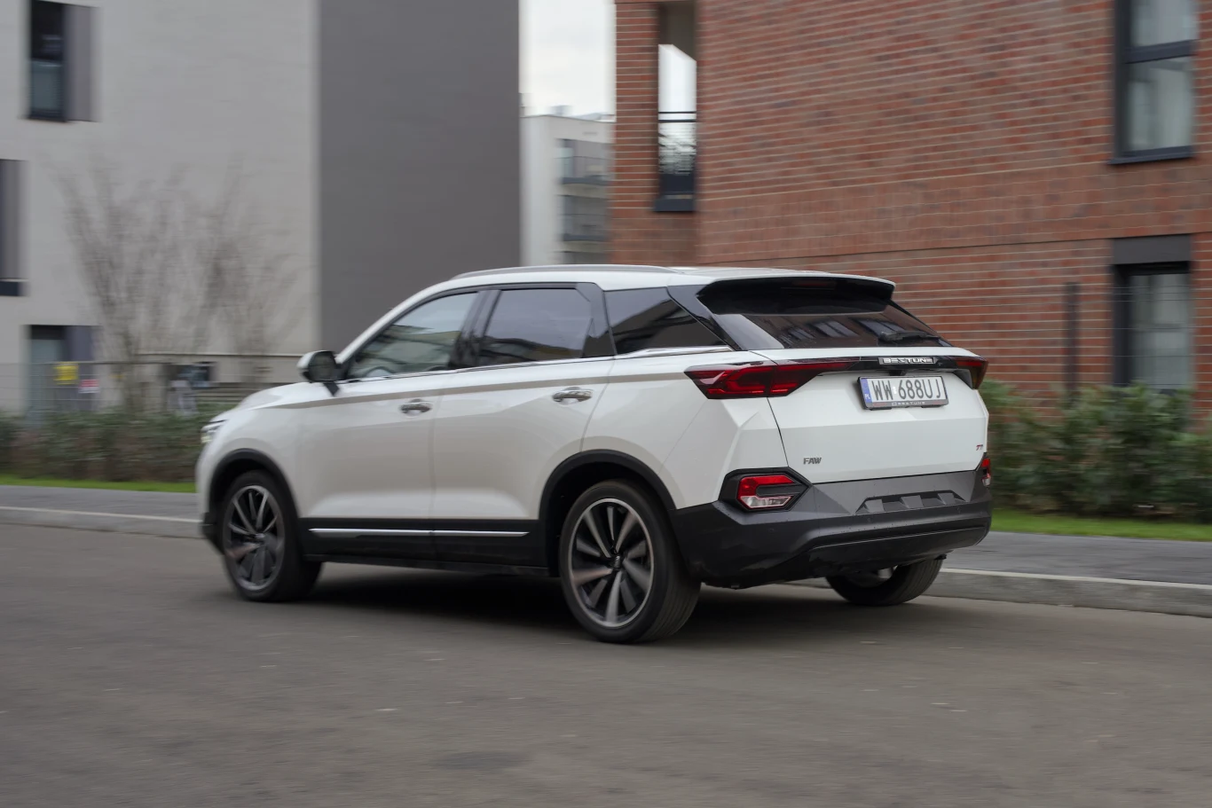 Biały SUV zaparkowany na ulicy, w tle nowoczesne budynki mieszkalne oraz niska roślinność, samochód stoi tyłem do aparatu.