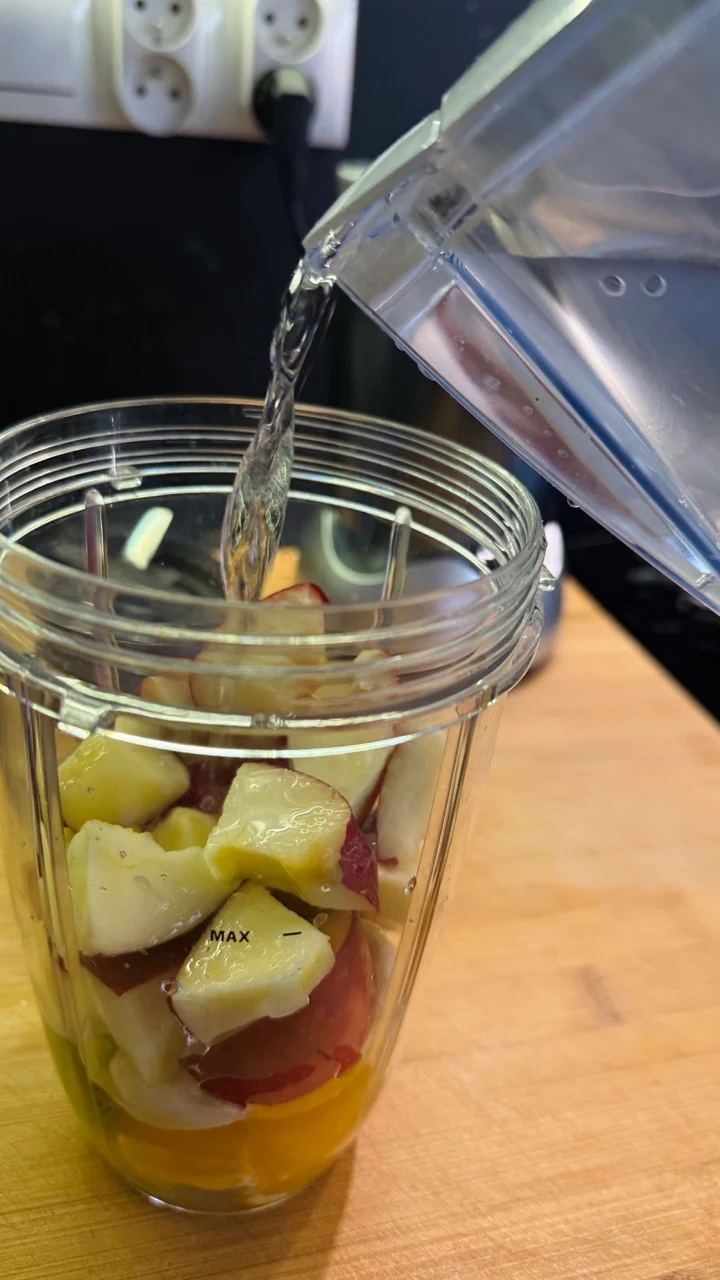 Koktajler z owocami takimi jak jabłka i brzoskwinie oraz kawałkiem pomarańczy, do którego wlewana jest woda z plastikowego dzbanka, całość stoi na drewnianym blacie kuchennym.