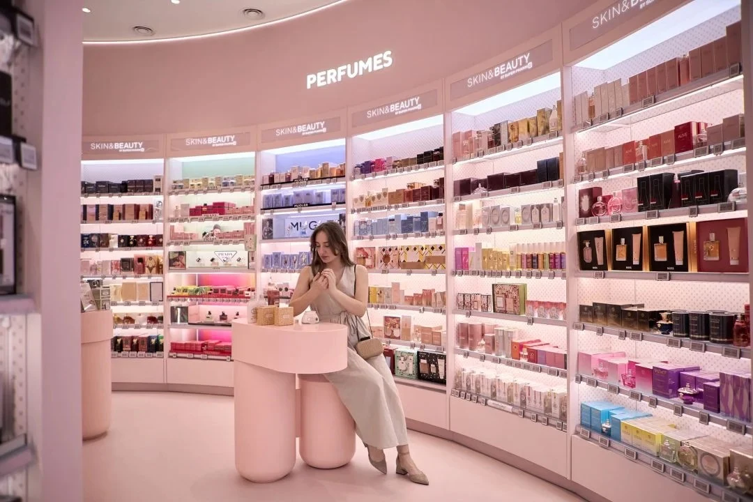 Elegancka kobieta siedzi przy okrągłym różowym stoliku w nowoczesnej perfumerii otoczonej jasnym wystrojem wnętrza, na półkach ustawione są liczne flakony perfum i kosmetyków, tworząc uporządkowaną i luksusową atmosferę.