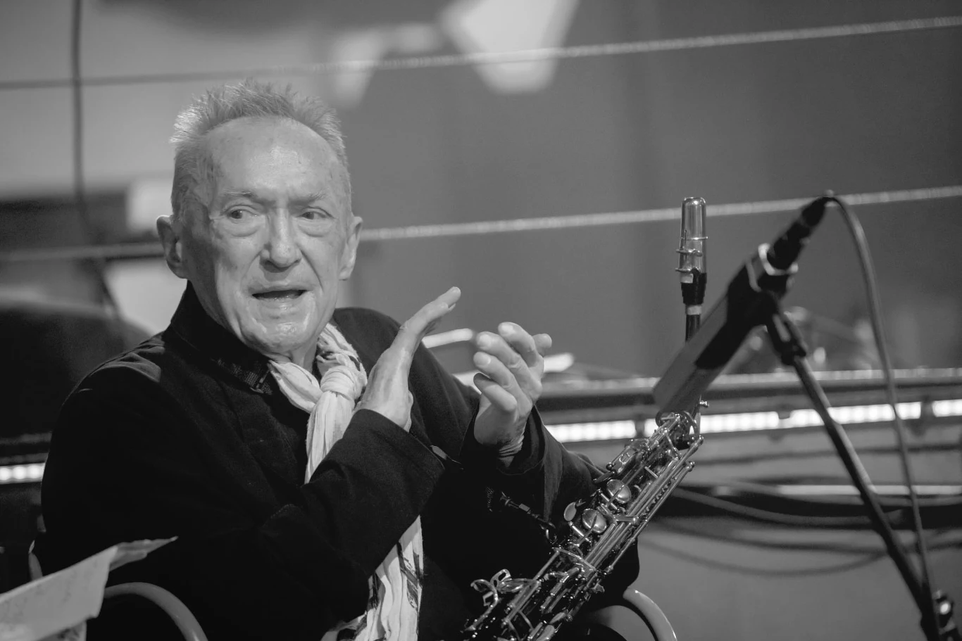 Michał Urbaniak zmarł w wieku 82 lat Starszy mężczyzna z szalikiem na szyi siedzi na scenie przy mikrofonie, obok niego saksofon. Wyraz twarzy skupiony i zaangażowany, w tle elementy sprzętu muzycznego.