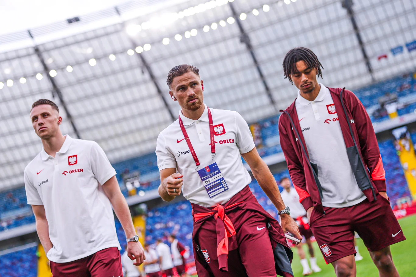 Marcin Bułka, Matty Cash i Maxi Oyedele Trzech piłkarzy w oficjalnych strojach reprezentacji Polski spaceruje po stadionie, tło stanowią zadaszone trybuny z niebieskimi krzesełkami. Każdy z zawodników nosi białą koszulkę polo z logiem Orlen i elementami czerwieni oraz sportowe spodnie.