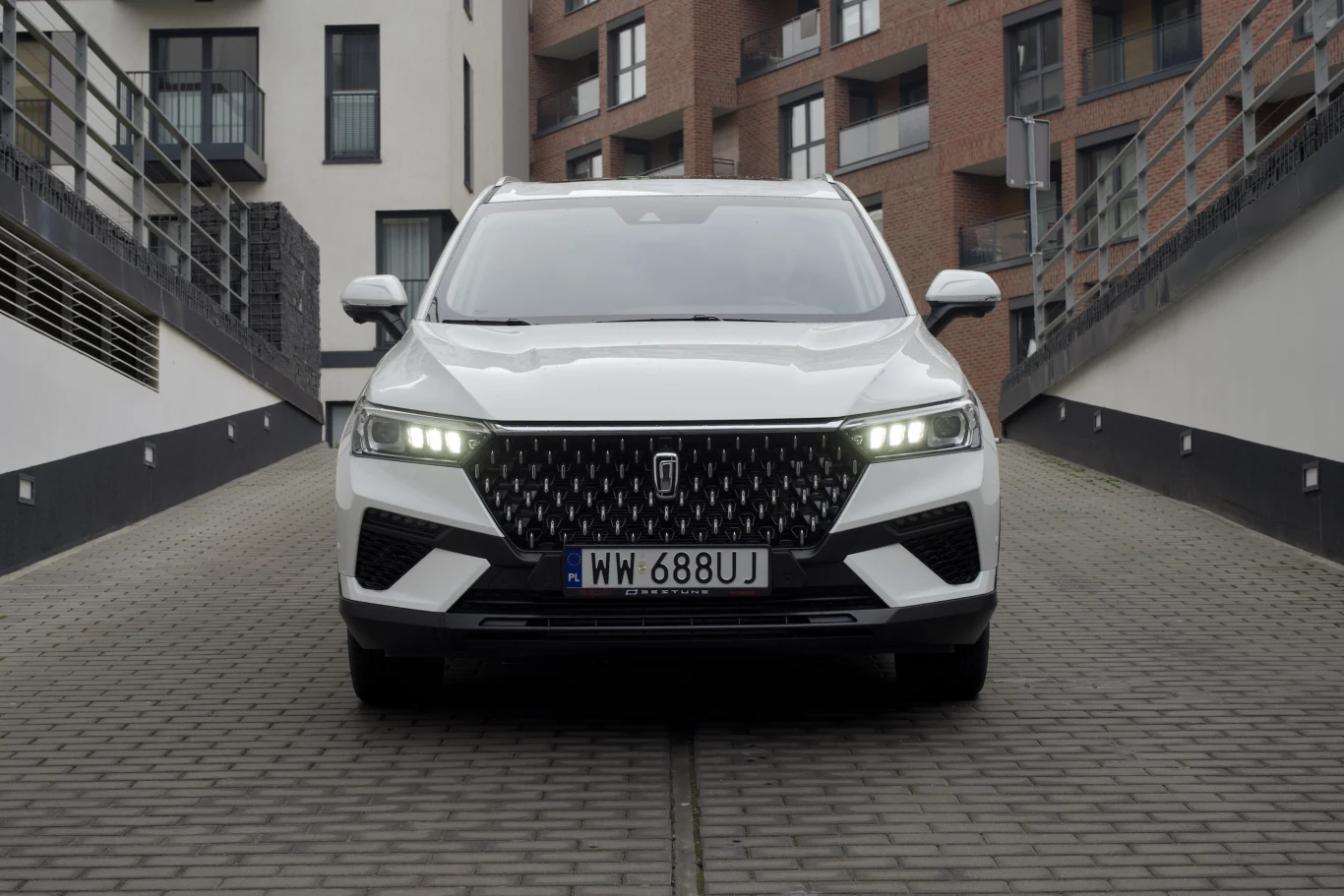 Biały nowoczesny SUV zaparkowany na brukowanej drodze pomiędzy nowoczesnymi budynkami mieszkalnymi, z zapalonymi światłami dziennymi i dobrze widoczną rejestracją.