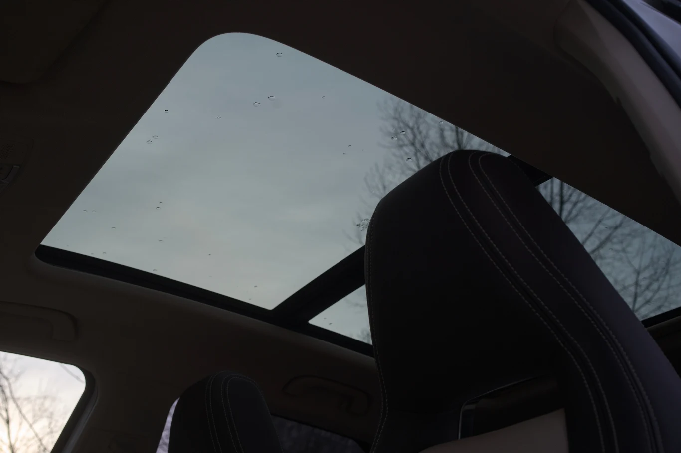 Panoramiczny, przeszklony dach samochodu z widokiem na niebo oraz fragmenty przednich foteli, lekko zarysowane kontury gałęzi drzew na zewnątrz.