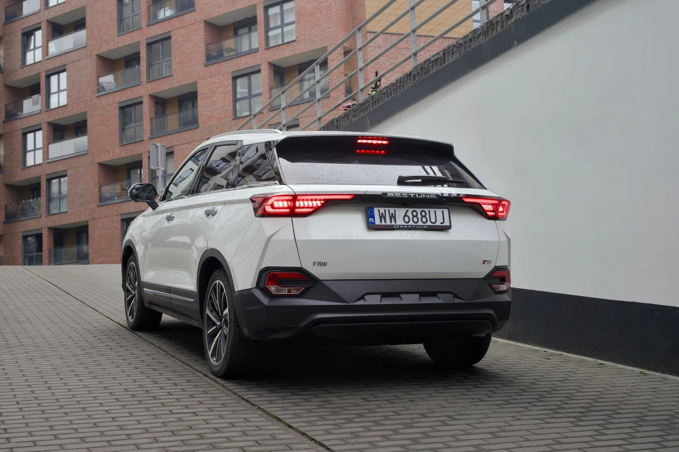 Biały SUV o nowoczesnych liniach, zaparkowany na pochylni przy budynku mieszkalnym z czerwonej cegły, charakteryzuje się wyrazistymi światłami tylnymi oraz widoczną rejestracją.