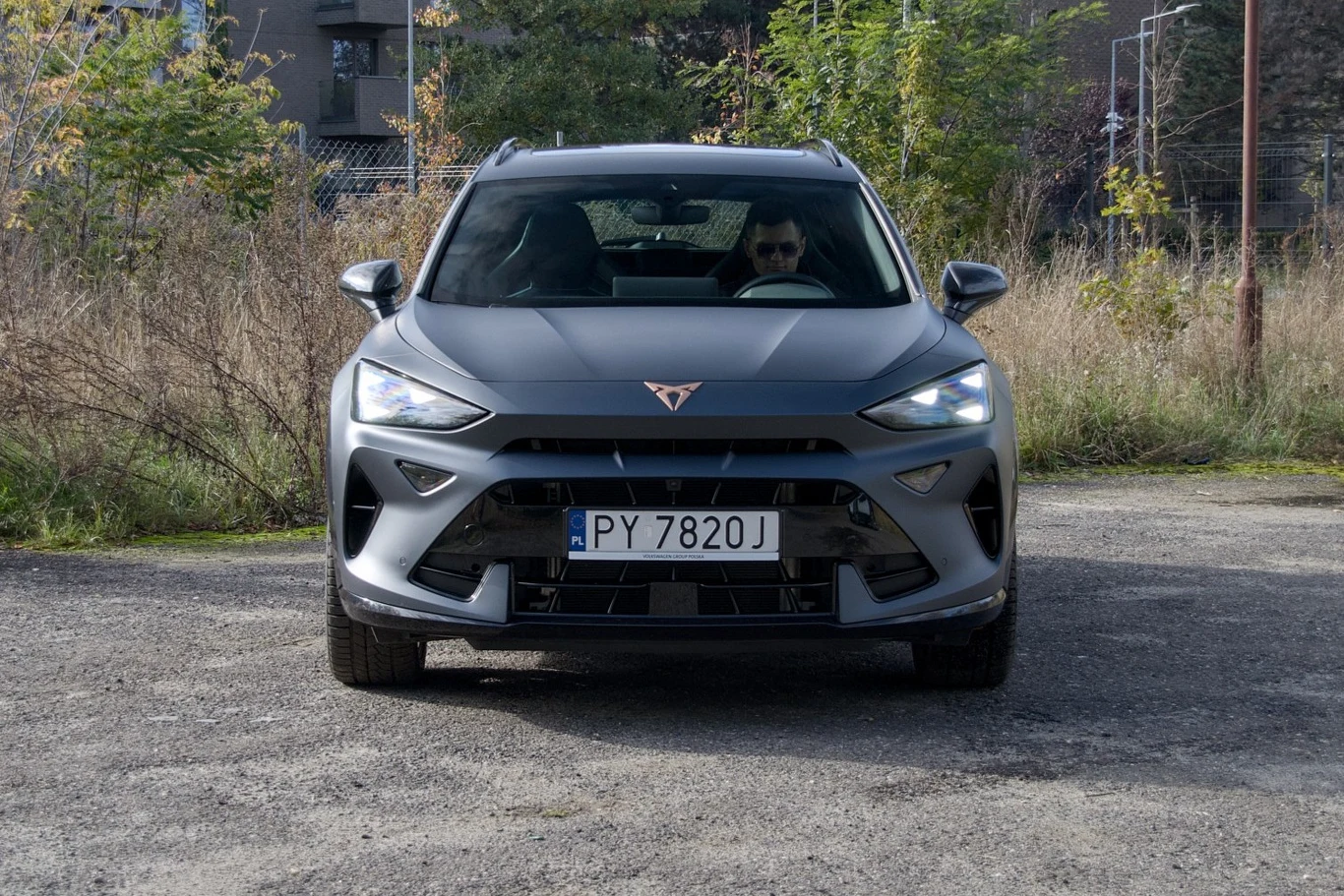 Nowoczesny, szary samochód Cupra Formentor widziany od przodu, na tle nieuporządkowanej miejskiej przestrzeni z roślinnością oraz budynkami w tle, za kierownicą widoczna osoba.