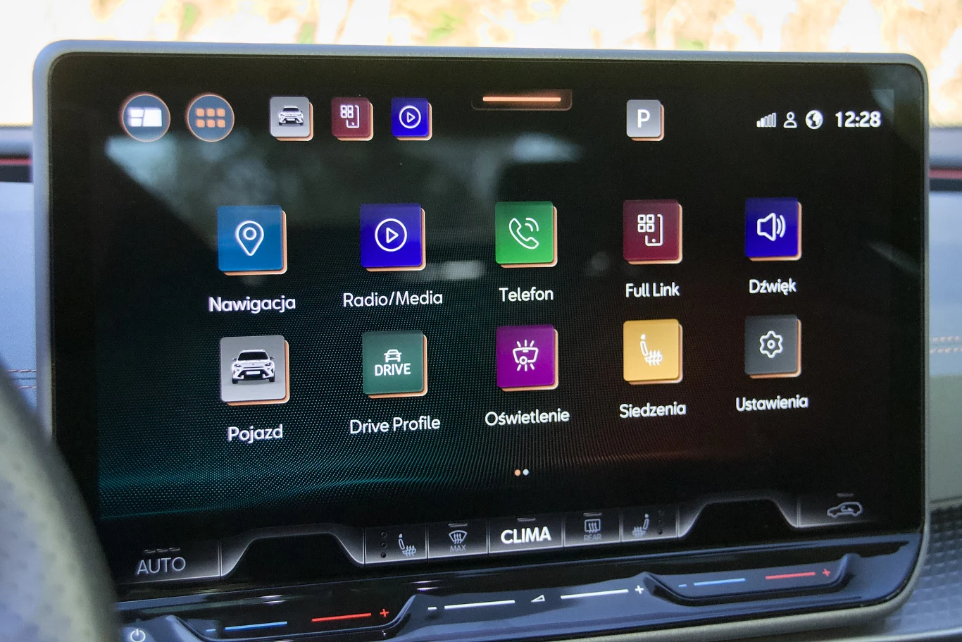 Kolorowy ekran dotykowy w samochodzie prezentujący menu systemu multimedialnego z ikonami dotyczącymi nawigacji, radia, telefonu, ustawień pojazdu, profilu jazdy, oświetlenia, siedzeń oraz innych funkcji.