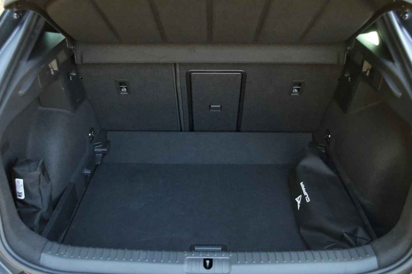 Pusty bagażnik samochodu z czarną tapicerką, z wyraźnie widocznymi oznaczeniami i organizerem po prawej stronie oraz składanym otworem w tylnej kanapie.