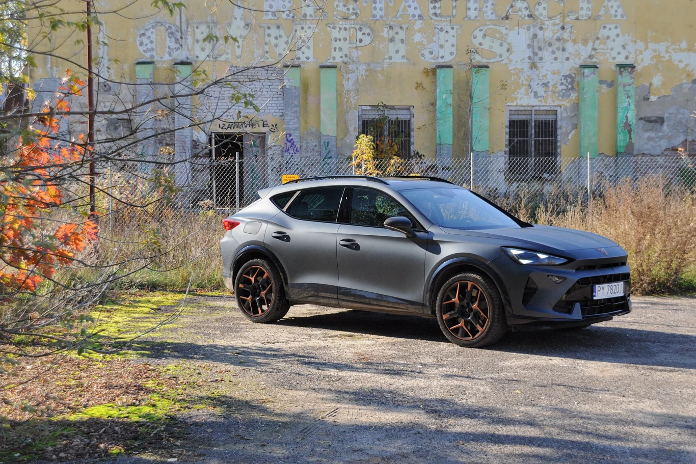 Nowoczesny samochód SUV w matowym, ciemnym kolorze zaparkowany przed zniszczonym, opuszczonym budynkiem z wyblakłym napisem Restauracja Campus. W tle wysoka, zardzewiała siatka i ślady degradacji oraz roślinność porastająca teren wokół.