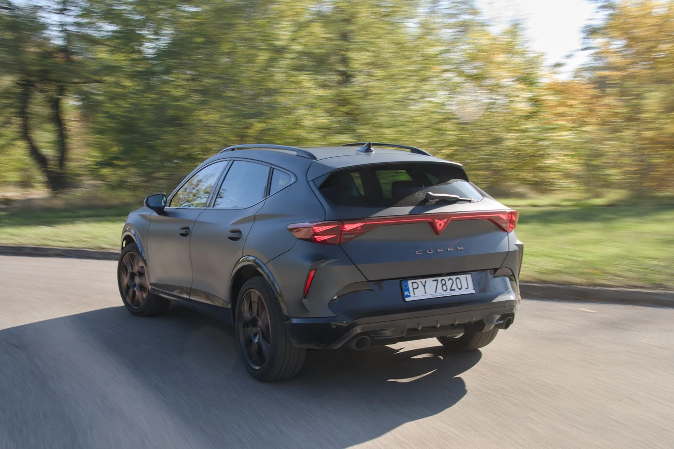 Czarny samochód typu SUV marki Cupra poruszający się po asfaltowej drodze, otoczony zielenią oraz drzewami w tle, rejestracja PY 7820J widoczna na tylnej klapie pojazdu.