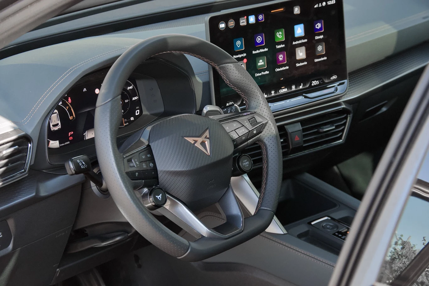 Kierownica z logo marki Cupra, zaawansowana deska rozdzielcza z cyfrowym zegarem, duży ekran dotykowy systemu multimedialnego z widocznymi ikonami aplikacji, nowoczesne wnętrze auta zaprojektowane z dbałością o detale.