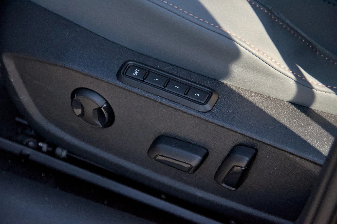 Panel boczny fotela samochodowego z widocznymi przyciskami do regulacji ustawień siedziska i zapamiętywania pozycji, wykonanie z czarnego plastiku oraz fragment tapicerki z dekoracyjnym przeszyciem.