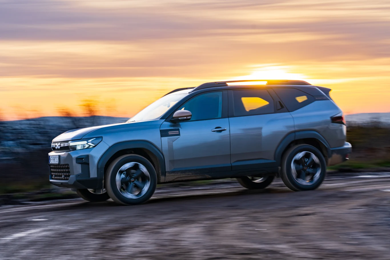 Nowoczesny SUV w kolorze szarym jadący po nieutwardzonej drodze o zachodzie słońca, z widocznymi odbiciami światła na nadwoziu, dynamicznie uchwycony w ruchu na tle horyzontu.