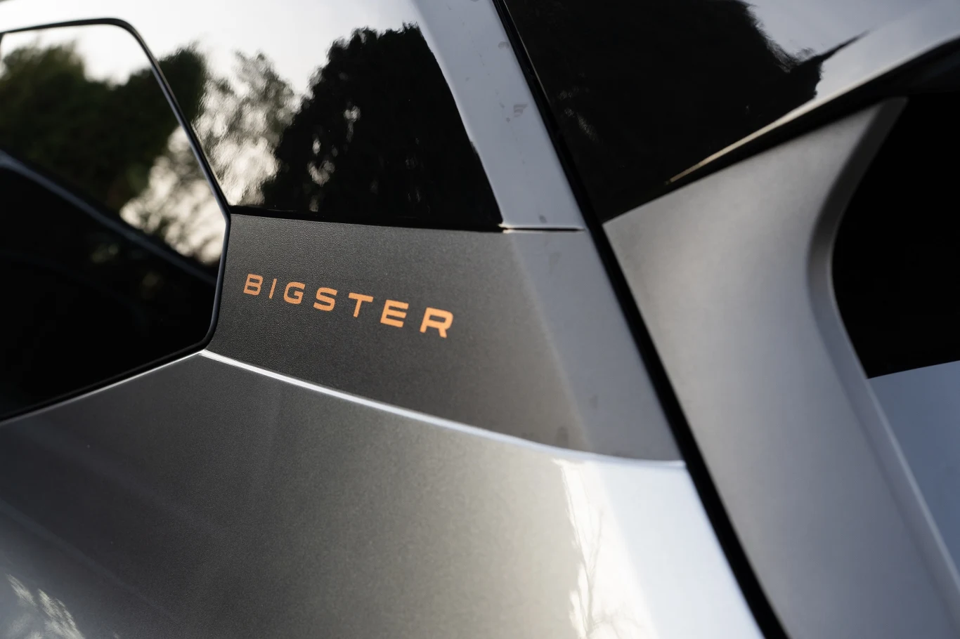 Detal karoserii auta w kolorze srebrnym z widocznym napisem BIGSTER na czarnej powierzchni przy oknie, w tle rozmazane drzewa odbijające się w lakierze.